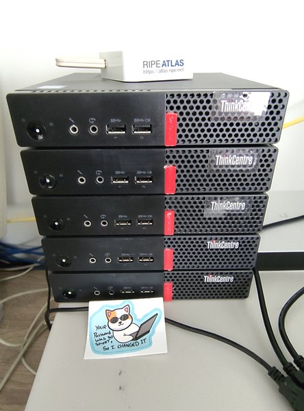 A photo of a stack of 5 Lenovo Thinkcentre Tiny M910q PCs on top of a Compaq Deskpro pIII ENS SFF. On top is a TP-Link based RIPE Atlas probe. There's a sticker with a handdrawn cat wearing sunglass, using laptop. A sentence reads 'your password was too short, so I changed it'.